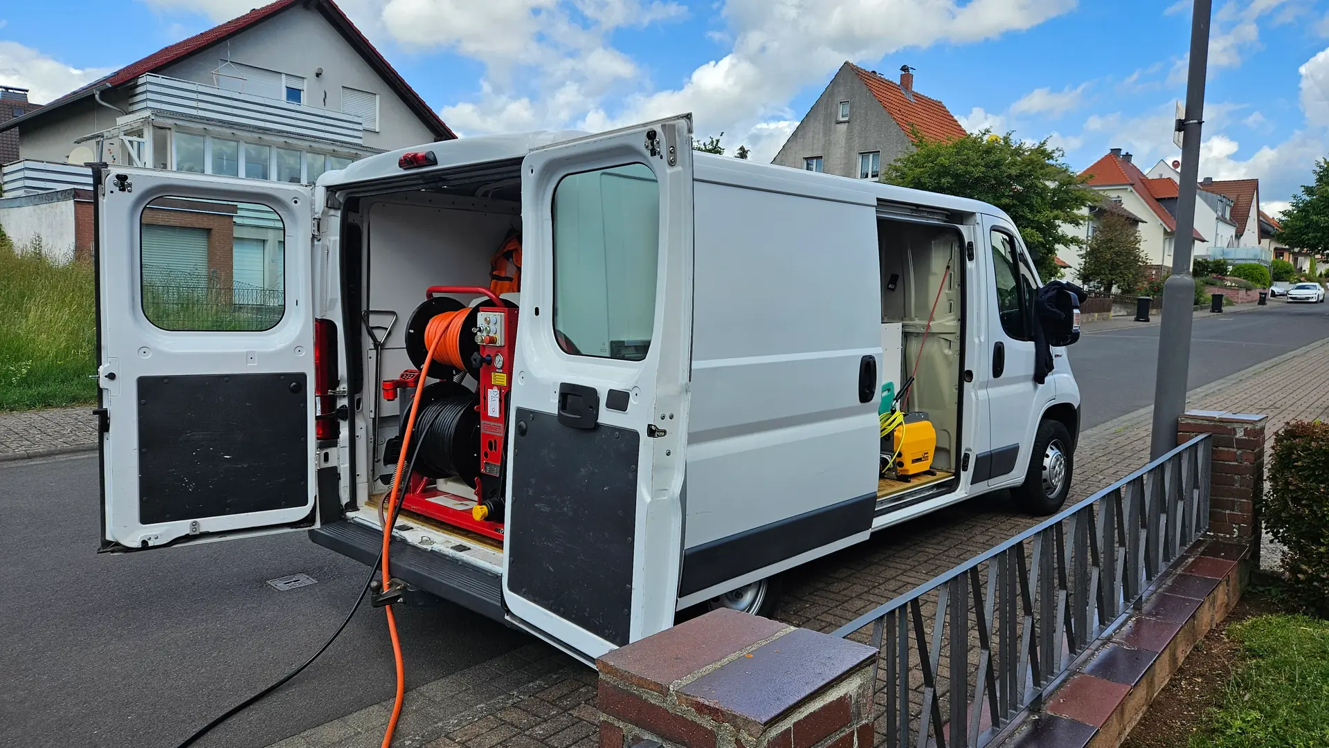 Firmenwagen der TK-Rohrreinigung Fulda mit geöffneten Türen und Spültechnik in einer Wohnstraße, der Schlauch führt zum Haus
